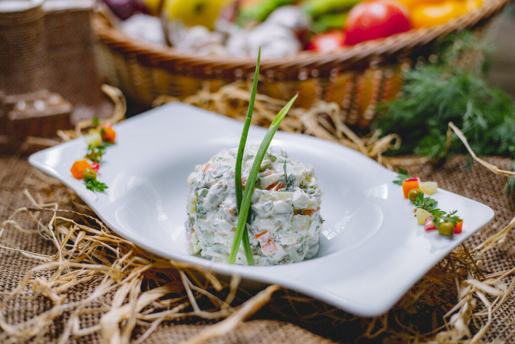 side view of traditional russian olivier salad with chicken decorated with green onion in a white bowl on straw background 1024x683